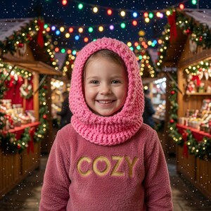 May include: A bright pink chunky knit balaclava hood worn over a dusty rose fleece sweatshirt featuring the word COZY embroidered in gold lettering across the chest. The garment is designed with a thick, textured yarn weave and a soft fleece fabric.
