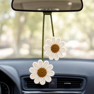 May include: A crochet car accessory featuring two white daisy flowers with brown centers, suspended from a dark olive green yarn cord. The flowers are attached at different lengths to the cord, which is designed to hang from a rearview mirror.