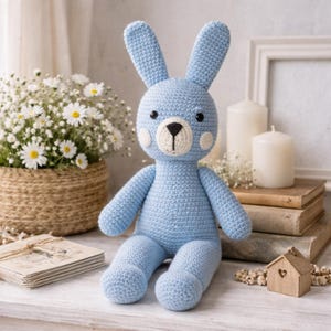 May include: A light blue crochet bunny with long ears, black eyes, and a white muzzle. The handmade stuffed animal has a round body and is sitting on a white surface. Daisies in a woven basket and candles are in the background.