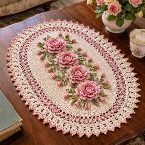May include: An oval crochet doily featuring a cream-colored base with a decorative scalloped border in shades of pink and cream. The center is adorned with four three-dimensional pink rose blossoms, green leaves, and small pink rosebuds.