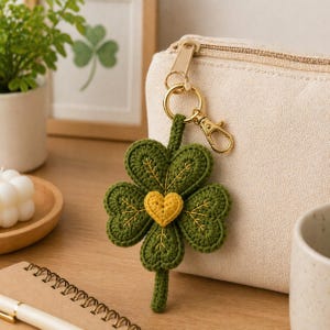 May include: A handmade green crochet four-leaf clover keychain with a yellow heart in the center. The clover is attached to a gold-colored clasp and ring. The keychain is hanging next to a cream-colored pouch.