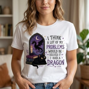 May include: A white t-shirt featuring a graphic of a black cat resting on a stack of books next to a lit candle and a flying dragon in a gothic window frame. The text reads: I THINK A LOT OF MY PROBLEMS WOULD BE SOLVED IF I HAD A DRAGON.