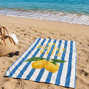 Peut inclure: Une serviette de plage rectangulaire à rayures verticales bleues et blanches, ornée d'un motif jaune de deux citrons avec des feuilles vertes et du texte jaune LA DOLCE VITA. Un sac fourre-tout en paille et des lunettes noires sont à côté.