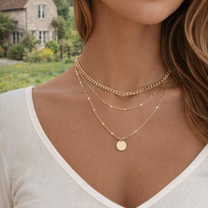 Peut inclure: Un collier doré à plusieurs rangs. La chaîne supérieure est une chaîne gourmette, la chaîne du milieu a de petits accents circulaires et la chaîne inférieure comporte un petit pendentif rond. Le collier est porté sur un haut blanc.