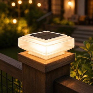 May include: A square solar post cap light with a translucent frosted glass body and a flat black solar panel on top. The light emits a warm glow from within, illuminating the edges of the fixture while mounted on a dark wooden fence post.