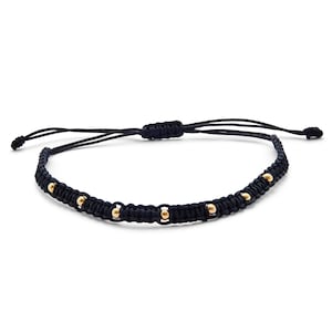 Könnte beinhalten: Schwarzes Makramee-Armband mit goldfarbenen Perlen. Das verstellbare Armband hat ein gewebtes Design mit gleichmäßig verteilten Metallperlen. Das Armband ist für das Handgelenk konzipiert.