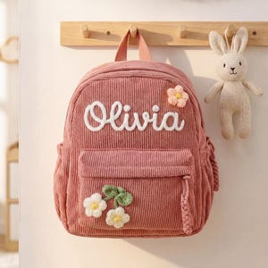 May include: A pink corduroy backpack with the name "Olivia" embroidered in white. The backpack has a front pocket, side pockets, and flower appliqués. A stuffed bunny is hanging nearby.