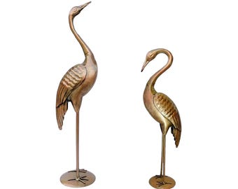 May include: Two standing crane sculptures crafted from metallic bronze-toned material. Each bird features a long neck, a pointed beak, and textured wing details. The figures are supported by thin metal legs attached to circular base plates.