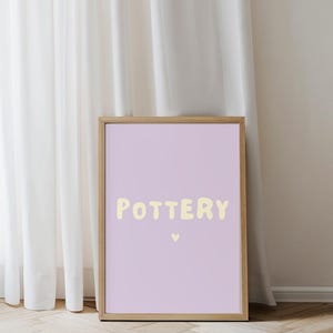 Könnte beinhalten: Ein gerahmter Druck mit dem Wort "POTTERY" in hellgelber Schrift auf einem lavendelfarbenen Hintergrund. Darunter befindet sich ein kleines Herz. Der Rahmen ist helles Holz. Der Druck lehnt an einem weißen Vorhang.