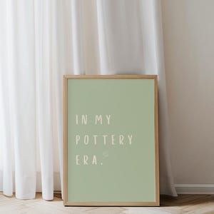 Könnte beinhalten: Ein gerahmter Kunstdruck mit dem Text "IN MY POTTERY ERA." in hellbeiger Schrift auf einem salbeigrünen Hintergrund. Der Rahmen besteht aus hellem Holz. Der Druck lehnt an einem weißen Vorhang und einer hellen Wand.