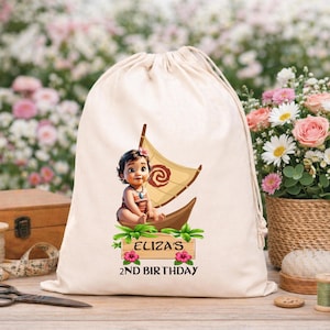 Puede incluir: Una bolsa beige con cord&oacute;n con una ilustraci&oacute;n de dibujos animados de un beb&eacute; en un barco con una vela. El texto "ELIZA'S 2ND BIRTHDAY" est&aacute; impreso debajo. La bolsa est&aacute; sobre una superficie de madera.