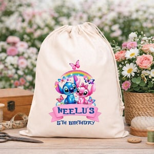 May include: A beige drawstring bag featuring a colorful graphic of two cartoon characters under a rainbow, surrounded by butterflies. The text "NEELU'S 5TH BIRTHDAY" is displayed on a pink banner. The bag is on a wooden surface.