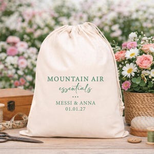 Könnte beinhalten: Ein cremefarbener Baumwollbeutel mit Kordelzug und dem grünen Aufdruck MOUNTAIN AIR essentials, den Namen MESSI & ANNA sowie dem Datum 01.01.27. Der Beutel hat einen gerafften Verschluss mit passenden Stoffkordeln und eine weiche Textur.