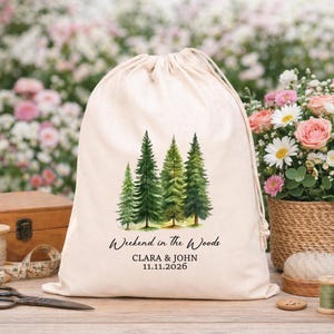 May include: A beige drawstring bag featuring a watercolor illustration of green pine trees. The text "Weekend in the Woods" is above the names "Clara & John" and the date "11.11.2026". The bag is sitting on a wooden surface.