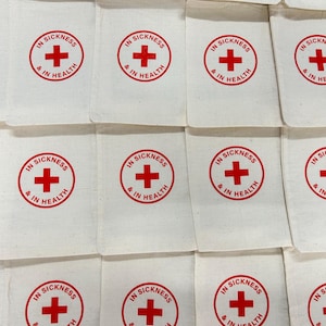 Puede incluir: Primer plano de peque&ntilde;as bolsas de tela blanquecina. Cada bolsa presenta una cruz roja dentro de un c&iacute;rculo rojo, con el texto "IN SICKNESS & IN HEALTH" en rojo. Las bolsas est&aacute;n dispuestas en un patr&oacute;n de cuadr&iacute;cula.