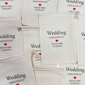 Puede incluir: Varios bolsos de tela blanquecina con cordones. Cada bolso tiene impreso en negro las palabras "Wedding HANGOVER KIT", un coraz&oacute;n rojo y los nombres "JESSI & TIMO" con la fecha "22.08.2025".