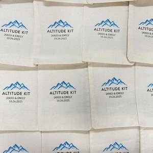 Puede incluir: Bolsas de tela blanquecina con un gr&aacute;fico de monta&ntilde;a azul y el texto "ALTITUDE KIT JARED & EMILY 10.26.2025". Las bolsas est&aacute;n dispuestas en un patr&oacute;n de cuadr&iacute;cula. El dise&ntilde;o se repite en varias bolsas.