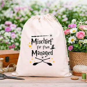 Peut inclure: Un sac en coton crème avec cordon de serrage, orné de l'inscription noire "Mischief far from Managed", d'une baguette magique, d'un chapeau de sorcière et de balais croisés. Des éclats d'étoiles dorées agrémentent le design. Le sac est sur une surface en bois.