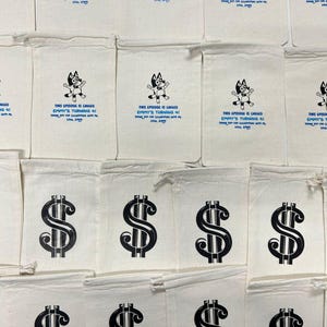 Puede incluir: Bolsas de lona blanquecinas con signos de d&oacute;lar negros impresos y texto. Algunas bolsas presentan un personaje de dibujos animados y el texto "THIS EPISODE IS CALLED EMMY'S TURNING 4!" en azul. Las bolsas est&aacute;n dispuestas en filas.