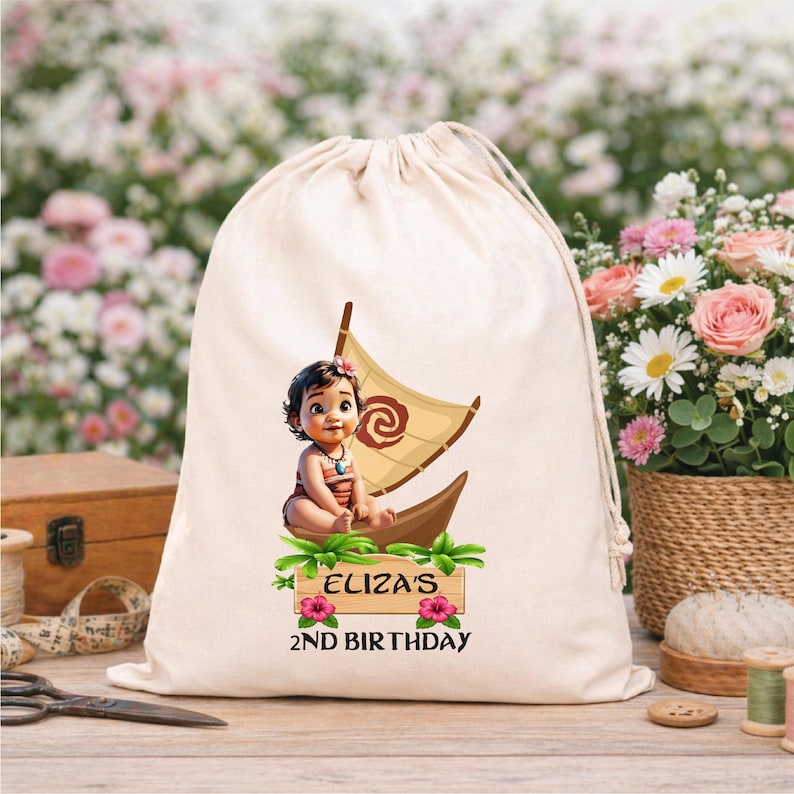 Puede incluir: Una bolsa beige con cord&oacute;n con una ilustraci&oacute;n de dibujos animados de un beb&eacute; en un barco con una vela. El texto "ELIZA'S 2ND BIRTHDAY" est&aacute; impreso debajo. La bolsa est&aacute; sobre una superficie de madera.