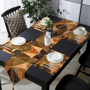 May include: A dining table set with a geometric patterned table runner and placemats in shades of brown and gold. White plates, gold and silver cutlery, and a small potted plant are arranged on the table. A mug with the number 12 is also visible.