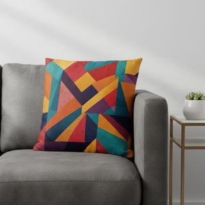 May include: A decorative throw pillow with a geometric design. The pillow features a colorful abstract pattern of triangles and other shapes in shades of red, orange, yellow, teal, and purple. The pillow is on a gray couch.