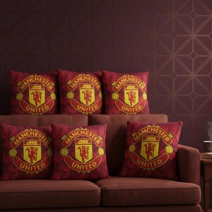 May include: A set of seven red decorative throw pillows with the Manchester United logo in yellow. The pillows are arranged on a brown sofa. The logo features a shield with a devil and the words "Manchester United".