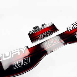 May include: A set of glossy adhesive decals for a Mercury 90 outboard motor. The decals feature a black and red gradient background with the white text MERCURY and the number 90. A smaller rectangular decal includes a stylised icon of a face.