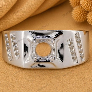 May include: A polished silver-tone ring featuring a central square-shaped setting designed to hold a single stone, flanked by two diagonal rows of small clear stones on each side. The band has a wide, smooth, and reflective metallic surface.