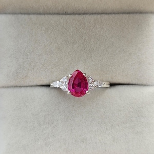 Könnte beinhalten: Ein silberner Ring mit einem großen, birnenförmigen, leuchtend roten Edelstein als Mittelpunkt. Flankiert wird der zentrale Stein von kleineren, klaren, dreieckig geschliffenen Akzentsteinen. Der Ring ist vor einem neutralen Hintergrund platziert.