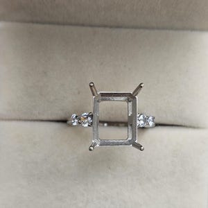 May include: A silver ring setting with a rectangular centre and small round accent stones on the band. The ring is displayed inside a grey jewellery box.