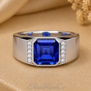 May include: A silver-toned ring featuring a large, square-cut, deep blue gemstone at the center. The band is flanked by two vertical rows of small, clear, round-cut stones on either side of the central blue gem. The text ElinaRoseCo is visible.