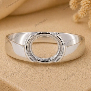 Könnte beinhalten: Ein silberner Ring mit einer kreisförmigen, leeren Fassung, die für einen Edelstein ausgelegt ist. Die Ringschiene ist glatt und poliert und besitzt eine metallische Oberfläche, die das Licht reflektiert. Die Fassung ist mittig platziert.