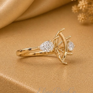 May include: A gold ring featuring a central marquise-shaped prong setting designed to hold a stone, flanked by a pear-cut clear stone on one side and a round-cut clear stone on the other, with a polished gold band and a delicate metal structure.