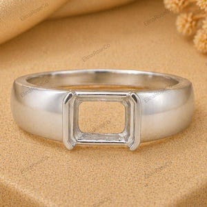 May include: A silver ring with a rectangular setting. The ring has a smooth, polished band and a square-shaped opening in the center, designed to hold a gemstone. The ring is displayed on a textured, tan surface.