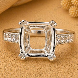 May include: A silver-toned ring setting featuring a rectangular prong setting designed to hold a center stone. The band is accented with a row of small, clear, round-cut stones embedded along each side of the shank, creating a polished look.
