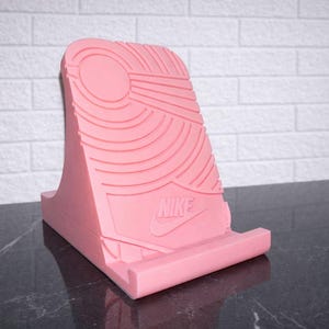 May include: A pink phone stand with a textured design resembling a shoe sole. The stand features the Nike logo and is designed to hold a phone or tablet. The stand is made of a solid material and is displayed on a dark surface.