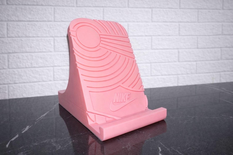 May include: A pink phone stand with a textured design resembling a shoe sole. The stand features the Nike logo and is designed to hold a phone or tablet. The stand is made of a solid material and is displayed on a dark surface.