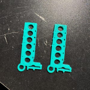 Può includere: Due componenti in plastica turchese stampati in 3D. Ogni pezzo ha una forma rettangolare con sei fori circolari e una piccola estensione arrotondata con un foro nella parte inferiore. Gli oggetti sono su una superficie scura.