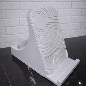 May include: A white, 3D-printed phone stand shaped like the sole of a sneaker. The stand features the Nike logo and detailed tread patterns. The stand is designed to hold a phone and is made of a solid, durable material.