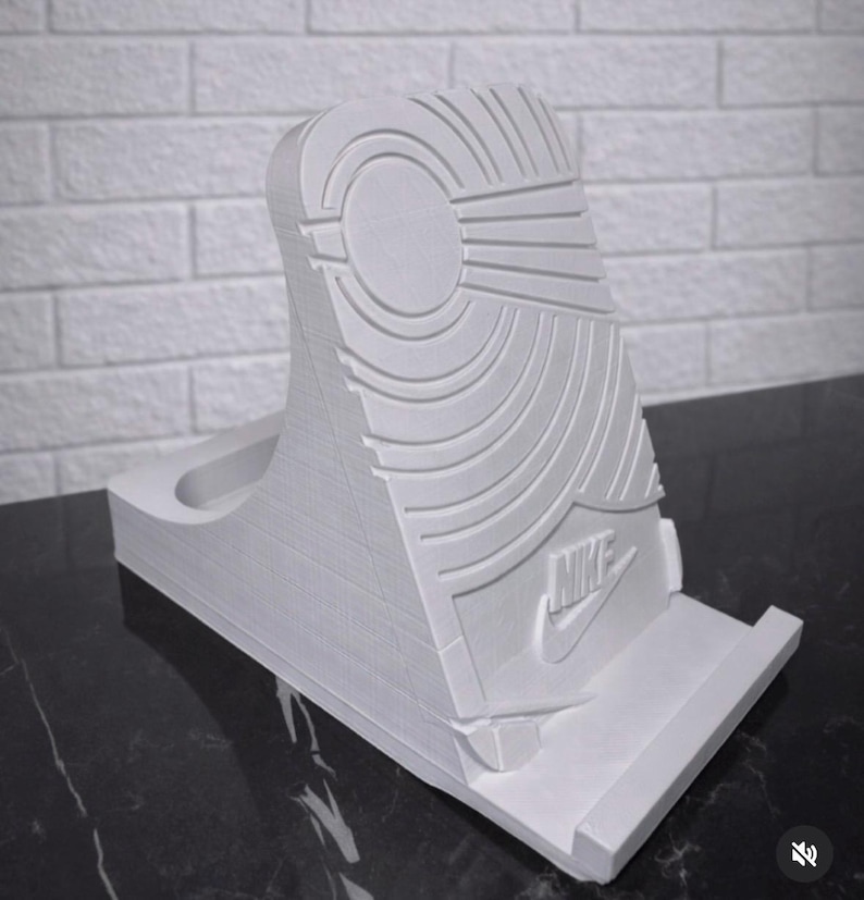 May include: A white, 3D-printed phone stand shaped like the sole of a sneaker. The stand features the Nike logo and detailed tread patterns. The stand is designed to hold a phone and is made of a solid, durable material.