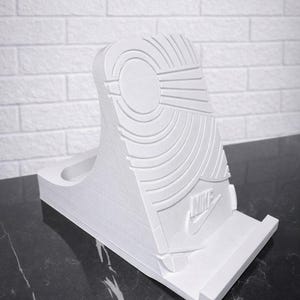 May include: White 3D-printed phone stand in the shape of a sneaker sole. The stand features the Nike logo and sole pattern. The stand is designed to hold a phone upright.