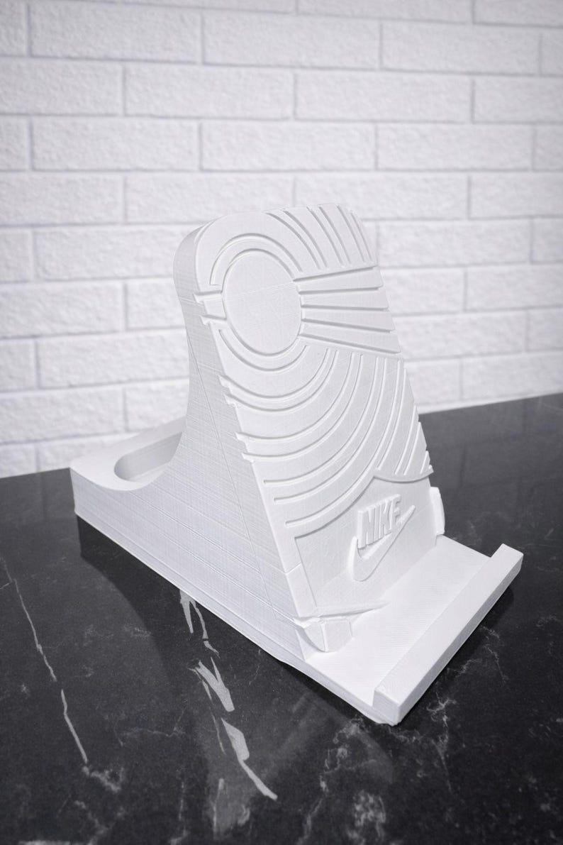 May include: White 3D-printed phone stand in the shape of a sneaker sole. The stand features the Nike logo and sole pattern. The stand is designed to hold a phone upright.