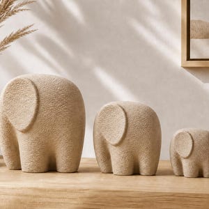 Puede incluir: Tres esculturas de elefantes beige de diferentes tamaños se exhiben sobre una superficie de madera. Los elefantes tienen cuerpos redondeados y orejas grandes y circulares. Las esculturas son de color neutro y tienen una superficie texturizada.
