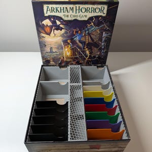May include: An Arkham Horror: The Card Game box with an open lid featuring dark fantasy artwork. The interior contains a grey 3D-printed organiser with a central honeycomb divider, several black card slots, and a series of colourful plastic dividers.