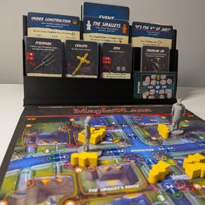 May include: A board game setup with a black card holder displaying cards labelled "UNDER CONSTRUCTION", "THE SMALLEYS", and "IT'S THE 4TH OF JULY". A game board with a map and yellow game pieces is in the foreground.