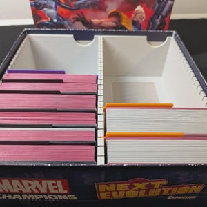 May include: An open Marvel Champions: The Card Game expansion box, labelled "Next Evolution." Inside, stacks of cards with pink and white edges are visible, separated by dividers. The box art displays comic book style illustrations.
