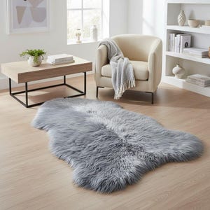 Puede incluir: Una alfombra gris de piel de oveja sintética con una textura suave y peluda se encuentra sobre un suelo de madera clara. La alfombra está en un salón, cerca de un sillón beige y una mesa de centro de madera con marco de metal negro.