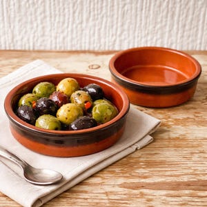 May include: Two terracotta bowls, one filled with assorted olives and a spoon, the other empty. The olives are green, black, and yellow, with red peppers. The bowls are on a beige napkin and a wooden surface.