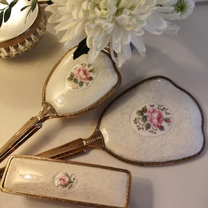 以下が含まれることがあります： ブラシ、手鏡、長方形の箱がセットになったヴィンテージの化粧台セット。クリーム色の背景に花柄のデザインと金色のトリムが施されています。各ピースには、ピンクのバラのイラストが描かれた中央の楕円形があります。